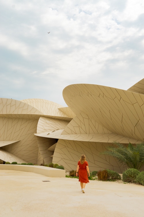 Een vrouw in een rode jurk loopt onder een bewolkte hemel richting de moderne, beige, bloemblaadjeachtige structuren van het Nationaal Museum van Qatar. De unieke architectuur van het gebouw contrasteert met het groen en woestijnlandschap eromheen.