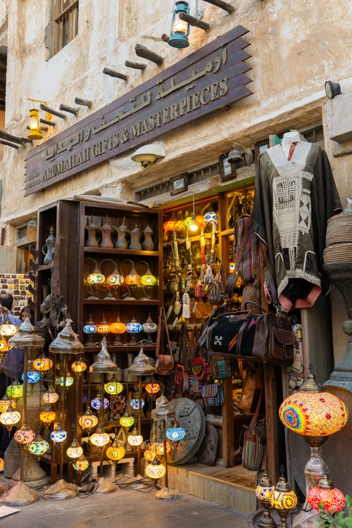 Een eclectische winkel in Souq Waqif, Doha, Qatar, met een levendige collectie kleurrijke mozaïeklampen, traditioneel aardewerk en geborduurde kledingstukken. De winkelpui, met het opschrift "ARUMAILIAH GIFTS & MASTERPIECES", is gevuld met een verscheidenheid aan handgemaakte ambachten en souvenirs onder warme, uitnodigende verlichting.