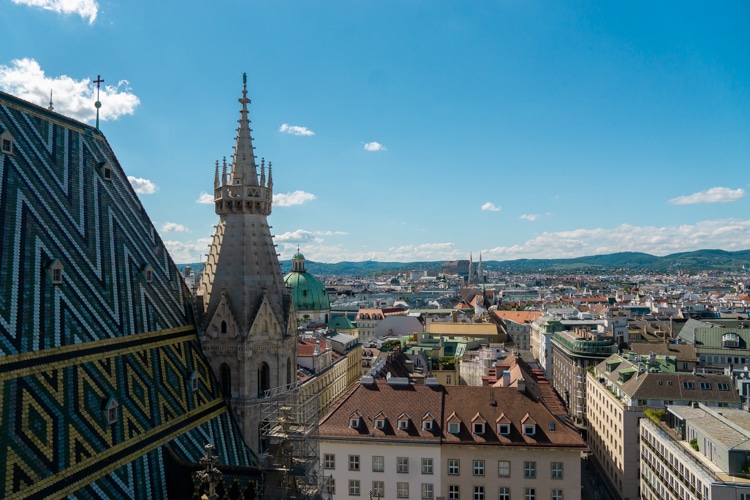 Uitzicht op Wenen vanaf de Stephansdom, met de gotische torenspits en het kleurrijke betegelde dak op de voorgrond, met een panorama van stadsgebouwen en verre heuvels onder een heldere blauwe lucht.