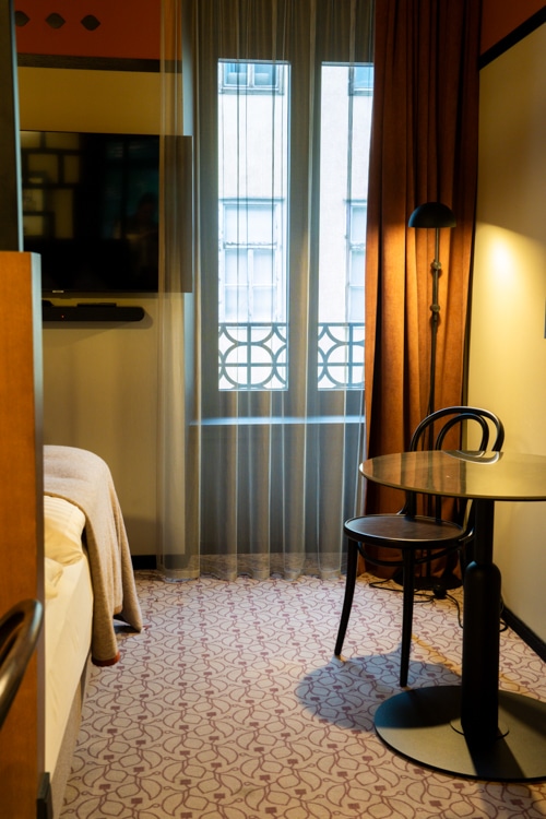 Gezellige hotelkamer met een bed, tv aan de muur, ronde tafel en twee stoelen naast een raam met vitrage en uitzicht op een aangrenzend gebouw.