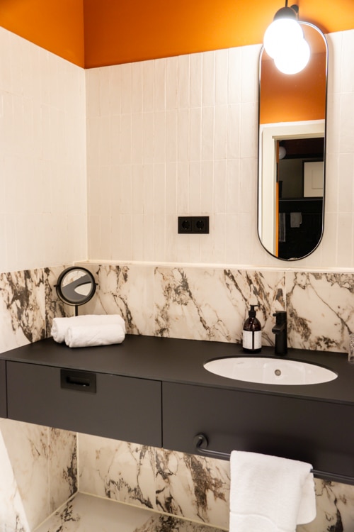 Moderne badkamer met een marmeren aanrechtblad, rechthoekige spiegel, oranje accentmuur en een witte handdoek netjes op het aanrecht naast toiletartikelen.