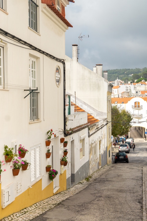 Een rustige straat in Sesimbra, met kleurrijke bloemen aan gevels en witte huizen langs een helling.