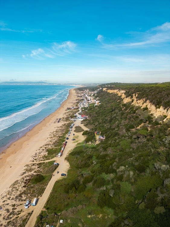 Luchtfoto van de Paisagem Protegida da Arriba Fóssil da Costa de Caparica, Portugal, met een uitgestrekt strand, golven die de kust raken, en huizen langs een onverharde weg