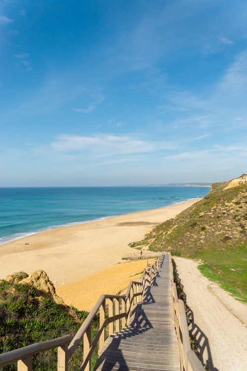 Houten pad dat leidt naar het uitgestrekte strand van Praia das Bicas in Sesimbra, Portugal, met helderblauw water en kliffen in de verte