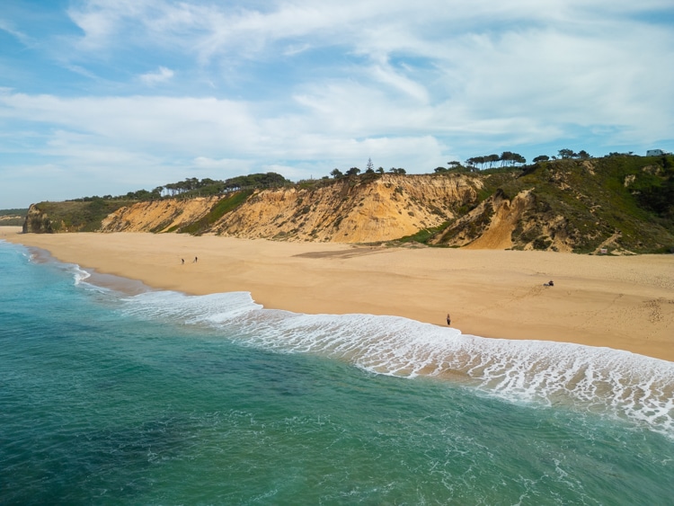 Luchtfoto van Praia das Bicas in Sesimbra, Portugal, met golven die het zandstrand raken, omringd door groene kliffen en een rustige omgeving
