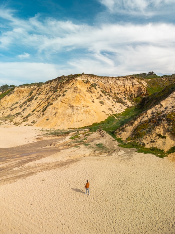 Breed zandstrand bij Praia das Bicas in Sesimbra, Portugal, met imposante gouden kliffen en een persoon die de uitgestrektheid van de natuur verkent onder een blauwe hemel