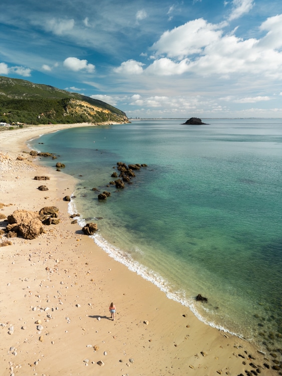 Een rustig strand in het natuurpark Serra da Arrábida, Portugal, met helder turquoise water, rotsen verspreid over het zand, en een klein eilandje in de verte