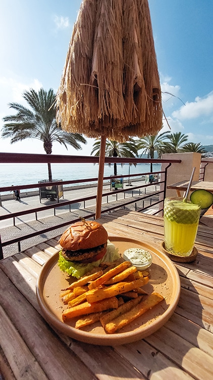 Een bord met een hamburger en frietjes, geserveerd met een verfrissend drankje op een terras met uitzicht op palmbomen en de zee in Sesimbra, Portugal, onder een rieten parasol.