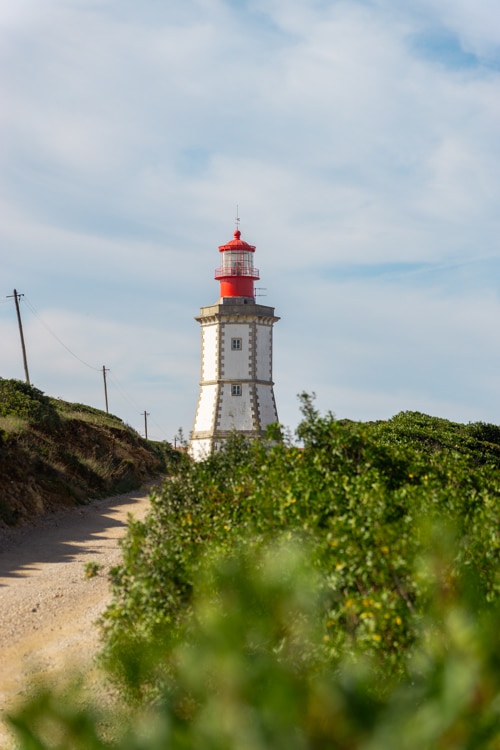 Vuurtoren van Cabo Espichel in Sesimbra, Portugal, gezien vanaf een onverhard pad omringd door groene struiken, met een heldere lucht op de achtergrond.
