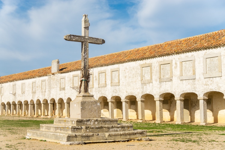Oude stenen kruis in de binnenplaats van het Santuário de Nossa Senhora do Cabo Espichel in Sesimbra, Portugal, met rijen van bogen en een rustieke uitstraling.