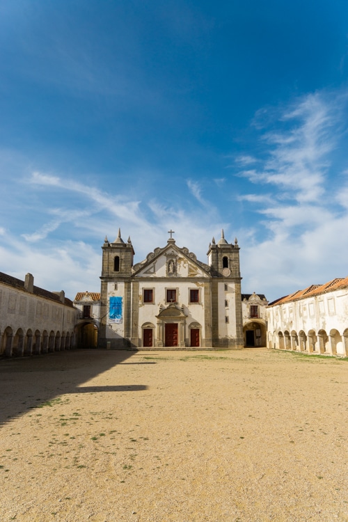 Voorzijde van het Santuário de Nossa Senhora do Cabo Espichel in Sesimbra, Portugal, met zijn historische architectuur en omliggende arcaden onder een heldere blauwe hemel.