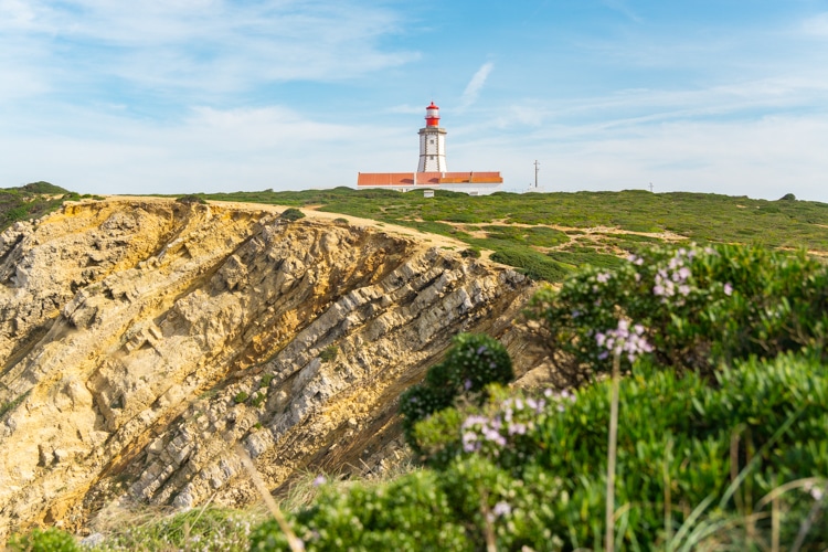 Uitzicht op de vuurtoren van Cabo Espichel in Sesimbra, Portugal, gelegen op een klif met dramatische rotsformaties en omgeven door groene vegetatie onder een heldere blauwe lucht.