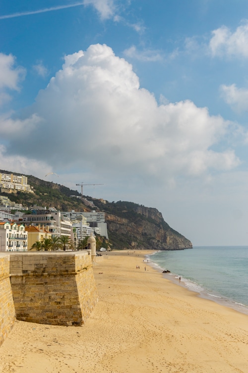 Uitzicht op het strand van Sesimbra, Portugal, met een historische stenen muur op de voorgrond. De kustlijn strekt zich uit langs helder zand en een rustige zee, met op de achtergrond groene heuvels en witte gebouwen. De blauwe lucht is versierd met grote, zachte wolken