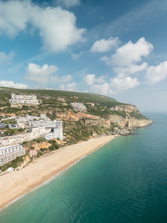 Uitzicht op Praia da Califórnia in Sesimbra, Portugal, met een gouden zandstrand, turquoise water en appartementencomplexen tegen de rotsachtige heuvels. 