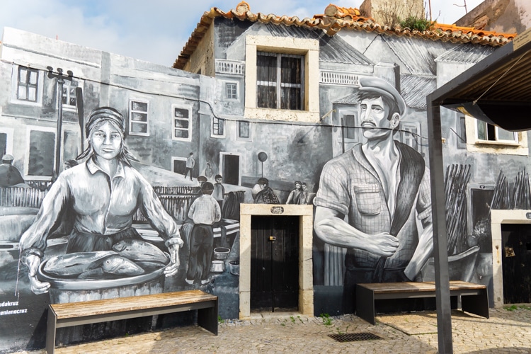 Straatkunst in Sesimbra, Portugal, met een zwart-wit muurschildering die vissers en een vrouw met een schaal vis afbeeldt, geplaatst op de gevel van een traditioneel gebouw met een oranje dak.