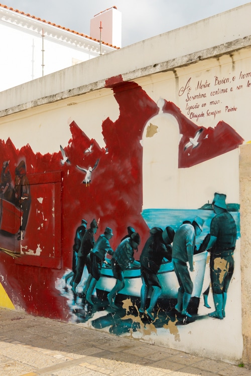Muurschildering in Sesimbra, Portugal, waarop vissers te zien zijn die samenwerken om een boot naar de kust te trekken, omringd door rode accenten en meeuwen