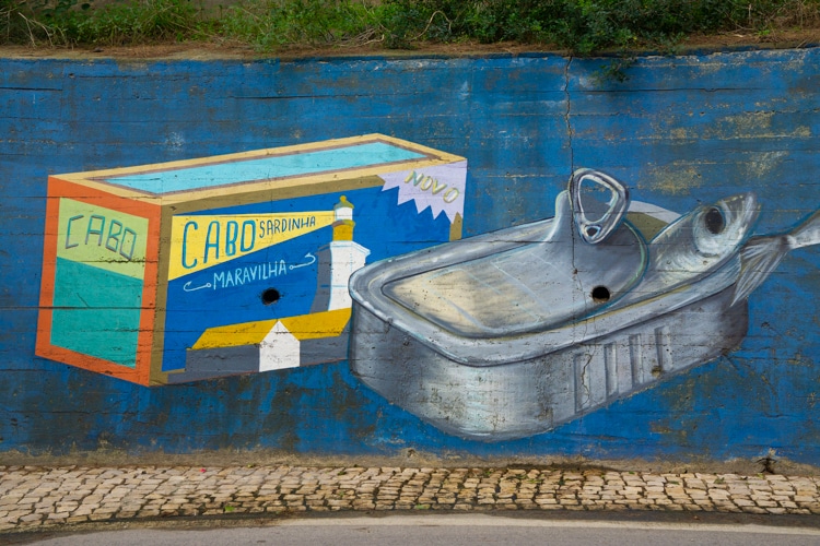 Muurschildering in Sesimbra, Portugal, met een kleurrijke sardineblik en een open blik met een sardine erin, tegen een blauwe achtergrond. 
