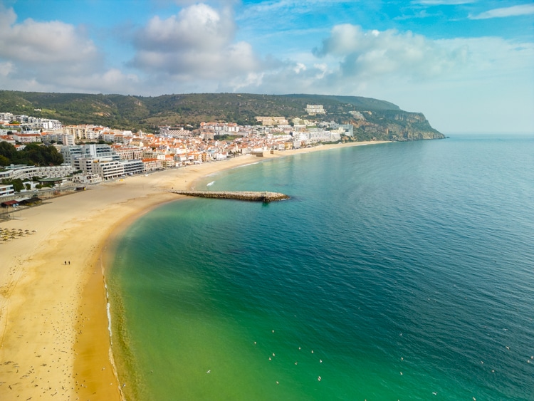Zicht op het strand van Sesimbra, Portugal, met helder turquoise water dat geleidelijk overgaat naar diepblauw. Op de achtergrond ligt de stad Sesimbra met haar witte gebouwen en oranje daken, tegen een achtergrond van groene heuvels. De lucht is licht bewolkt en de rustige zee nodigt uit voor een ontspannende stranddag.