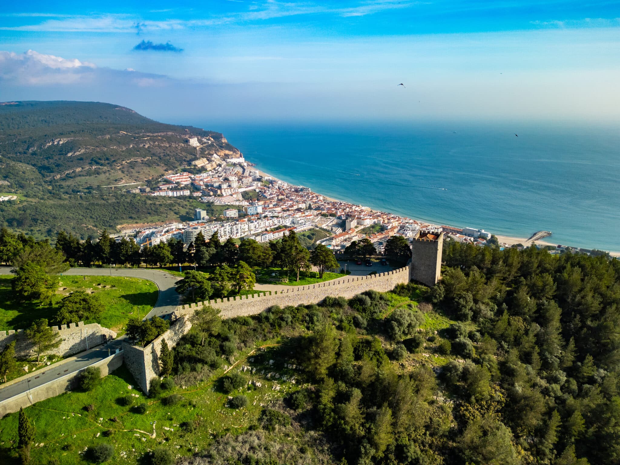 Uitzicht op de kustlijn van Sesimbra, Portugal, met op de voorgrond een middeleeuws kasteel dat zich uitstrekt over een groene heuveltop. De stad ligt aan de kust, met witte gebouwen en een strand dat uitkijkt over de blauwe Atlantische Oceaan. De omliggende heuvels en bossen creëren een schilderachtig contrast met de helderblauwe lucht en zee