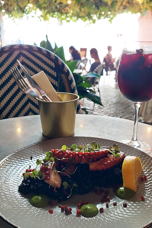 Een verfijnd gerecht van gegrilde octopus geserveerd op zwarte risotto, gegarneerd met microgroenten, een schijfje citroen en groene saus, bij Antu Alfama, een stijlvol restaurant in Lissabon. Op de achtergrond staat een glas rode sangria, wat perfect past bij de charmante ambiance van het restaurant, met zicht op een terras vol groen en gezellige zitjes.