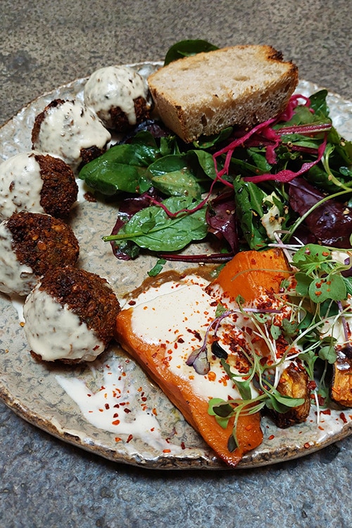 Een kleurrijk bord met falafelballetjes bedekt met tahinsaus, geroosterde groenten, een frisse salade en een stukje zuurdesembrood, geserveerd bij Honest Greens, een populair restaurant in Lissabon. Het gerecht straalt een gezonde en duurzame eetervaring uit, kenmerkend voor dit trendy restaurant.