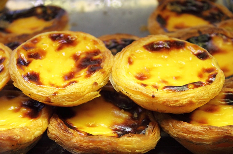 Een stapel versgebakken Pastéis de Belém, de iconische Portugese custardtaartjes met een krokante bladerdeegkorst en een romige vulling, licht gekarameliseerd aan de bovenkant. Deze wereldberoemde lekkernij is onmisbaar bij een bezoek aan Lissabon en wordt traditioneel geserveerd in de bakkerij van Pastéis de Belém.