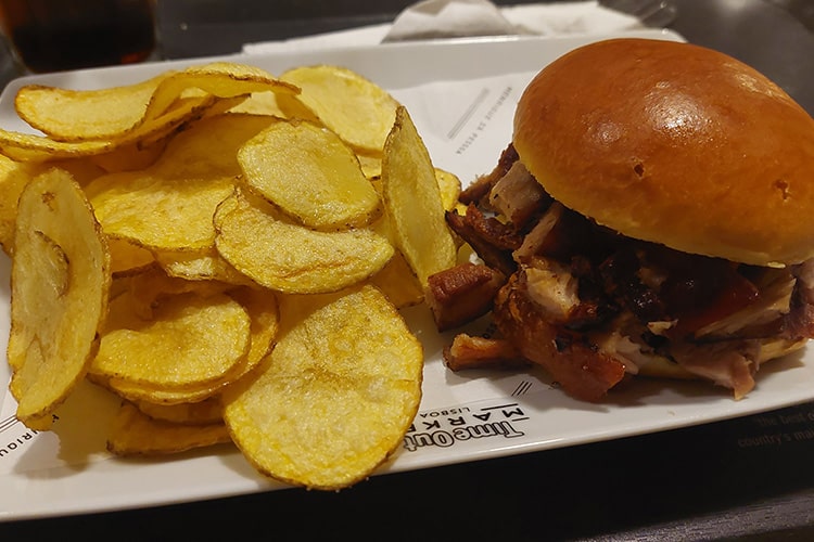 Een bord met een zacht broodje gevuld met sappig, gegrild vlees, geserveerd met een royale portie goudgele chips op een witte schaal. Het eten ziet er vers en smakelijk uit, perfect voor een gezellige maaltijd.