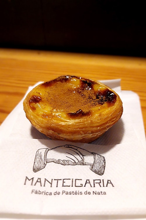 Een vers gebakken Pastel de Nata met een goudbruine, gekarameliseerde bovenkant, gepresenteerd op een servet met het logo van Manteigaria, een bekende fabriek voor deze traditionele Portugese custardtaartjes. De subtiele kaneel en het bladerdeeg zorgen voor een uitnodigende uitstraling.