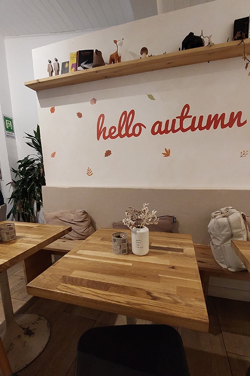 Een gezellige zithoek bij Quase Café, een van de sfeervolle restaurants in Lissabon, met houten tafels, beige kussens en een muurdecoratie met de tekst 'hello autumn', omringd door herfstbladeren. De warme en uitnodigende sfeer weerspiegelt de ontspannen ambiance van het café.