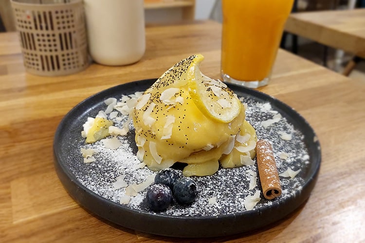Een bord met een decadent ontbijt bij Quase Café, een populair restaurant in Lissabon, bestaande uit pannenkoeken overgoten met citroenroomsaus, gegarneerd met kokosschilfers, blauwe bessen, een kaneelstokje en een schijfje citroen. Op de achtergrond staat een glas verse jus d’orange op een houten tafel, passend bij de smaakvolle presentatie van dit café-restaurant.