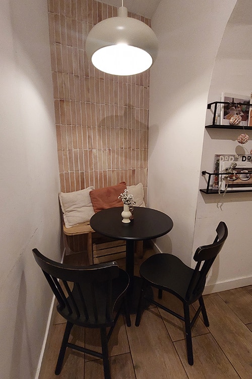 Een intieme zithoek in Quase Café, een charmant restaurant in Lissabon, met een ronde zwarte tafel, twee zwarte stoelen en een houten bankje met zachte kussens. De moderne witte hanglamp en een vaasje met gedroogde bloemen voegen een minimalistisch en sfeervol tintje toe aan de inrichting, typisch voor de beste restaurants in Lissabon