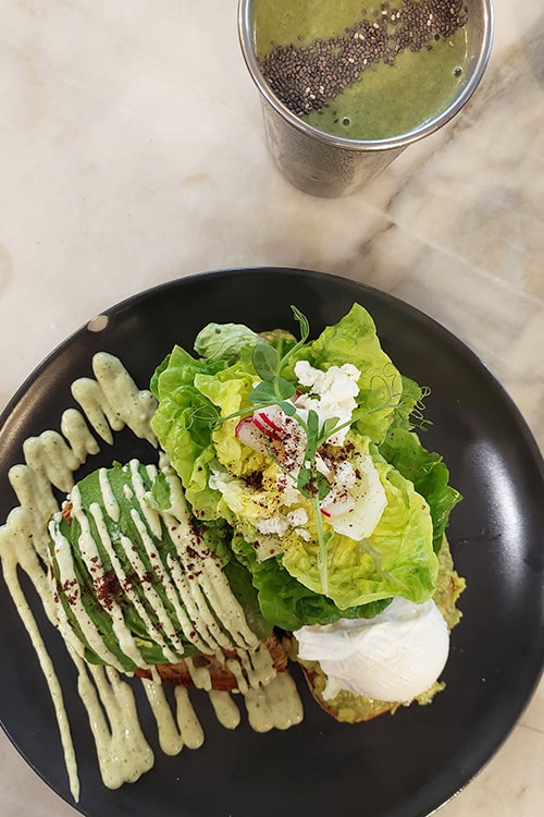 Een prachtig gepresenteerd ontbijt bij Dear Breakfast, een populair restaurant in Lissabon, met een bord gevuld met avocado op toast, romige groene saus, verse sla, een gepocheerd ei en decoratieve radijsjes. Naast het gerecht staat een smoothie met chiazaad, wat de focus op gezonde en verfijnde gerechten benadrukt die kenmerkend zijn voor dit trendy restaurant in Lissabon.