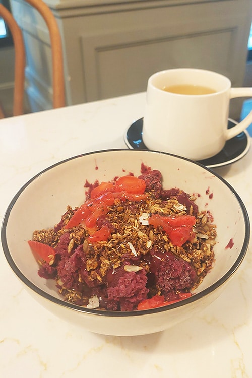 Een kleurrijke açai bowl geserveerd bij Hygge Kaffe, een knus restaurant in Lissabon, met een mix van romige açai, krokante granola, verse aardbeien en kokossnippers. Op de achtergrond staat een kopje warme thee, wat de rustgevende en gezellige sfeer van dit trendy café-restaurant benadrukt.