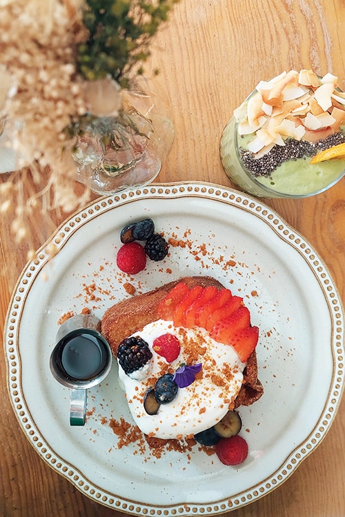 Een prachtig opgemaakte French toast geserveerd bij Miolo in Belém, een stijlvol restaurant in Lissabon, gegarneerd met verse aardbeien, bosbessen, frambozen, een toef slagroom en een vleugje kruimeldeeg. Ernaast staat een klein kannetje siroop voor extra zoetheid en een glas groene smoothie met kokosschilfers en chiazaad, wat de balans tussen indulgentie en gezondheid in dit restaurant benadrukt.