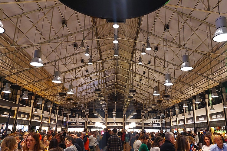 innenzicht van een drukke markthal in Lissabon met hoge, industriële plafonds en hangende lampen. Mensen genieten van eten en drinken aan tafels, omringd door kraampjes met diverse gerechten en dranken.