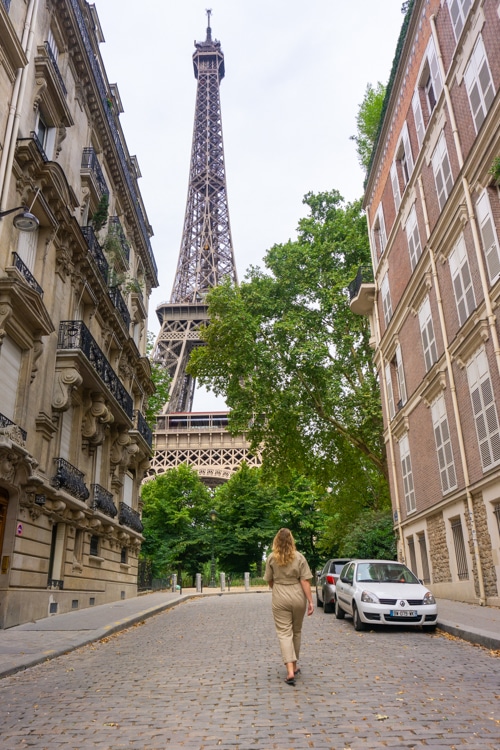 Een vrouw loopt over een geplaveide straat met een uitzicht op de Eiffeltoren, omringd door klassieke Parijse gebouwen. De foto vangt de elegantie en de betovering van een soloreis naar Parijs.