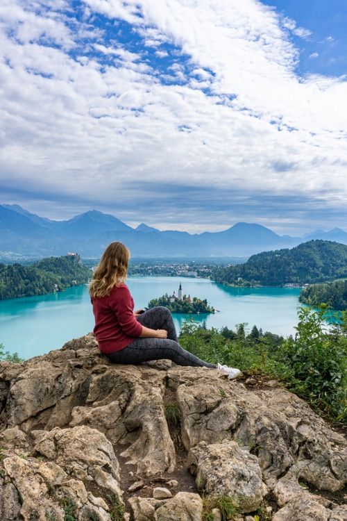 en vrouw zit op een rots en geniet van het panoramische uitzicht op het meer van Bled, met zijn beroemde eiland en kerkje, omringd door groene heuvels en bergen. De rust en schoonheid van dit moment benadrukken het plezier van soloreizen.