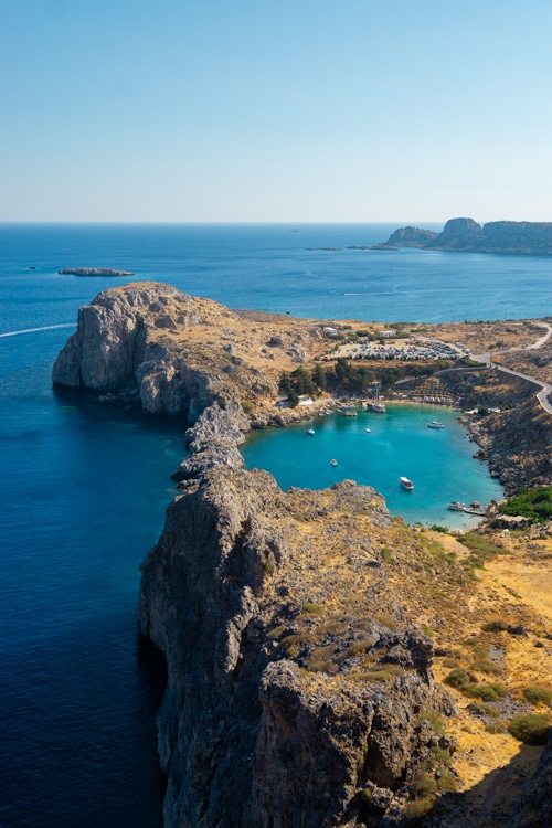 Een luchtfoto van de iconische St. Paul's Bay bij de Akropolis van Lindos op Rhodos, met turquoise water dat wordt omringd door steile kliffen en een smal strand. Kleine boten dobberen in het heldere water, terwijl een kronkelende weg de kustlijn volgt.