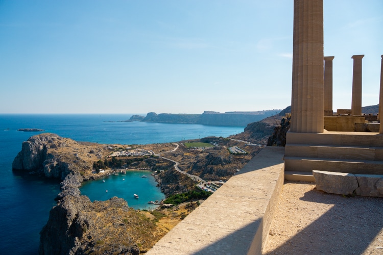 De afbeelding toont een panoramisch uitzicht op de Akropolis van Lindos op Rhodos, met op de voorgrond oude Dorische zuilen en in de verte een helderblauwe baai omringd door ruige kliffen en een historische nederzetting. Het heldere weer benadrukt de schoonheid van de omgeving, terwijl de zon schaduwen werpt op de oude stenen structuur.