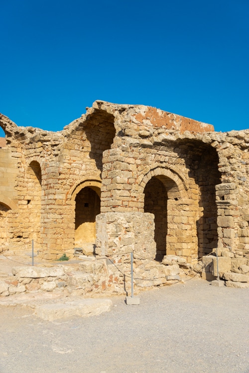 De ruïnes van een stenen gebouw op de Akropolis van Lindos, met bogen en verweerde muren die de sporen van eeuwen geschiedenis tonen. Het warme, okergele steen contrasteert prachtig met de heldere blauwe lucht erboven.