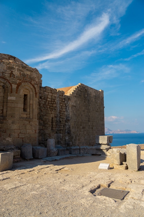 Een oud stenen gebouw met verweerde muren op de Akropolis van Lindos, met losse bouwstenen verspreid over de grond. De achtergrond toont een prachtig uitzicht op de zee en een heldere lucht, wat de historische sfeer aanvult.