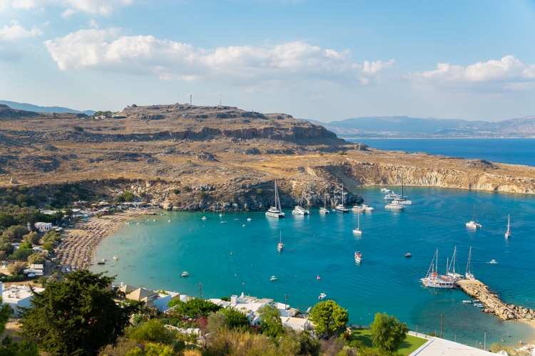 Een levendige baai vol met zeilboten en jachten, omringd door heuvels en een klein strand met parasols en witte huisjes. De zachte golven van de turquoise zee vormen een rustgevende aanvulling op de zonnige, mediterrane sfeer.