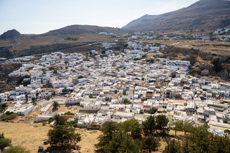 Een panoramisch uitzicht op het dorp Lindos, met een dicht netwerk van witgekalkte huizen die contrasteren met de dorre heuvels en bergen in de achtergrond. De charmante, traditionele architectuur wordt omringd door groen en kronkelige wegen, wat de rustieke sfeer van het dorp benadrukt.