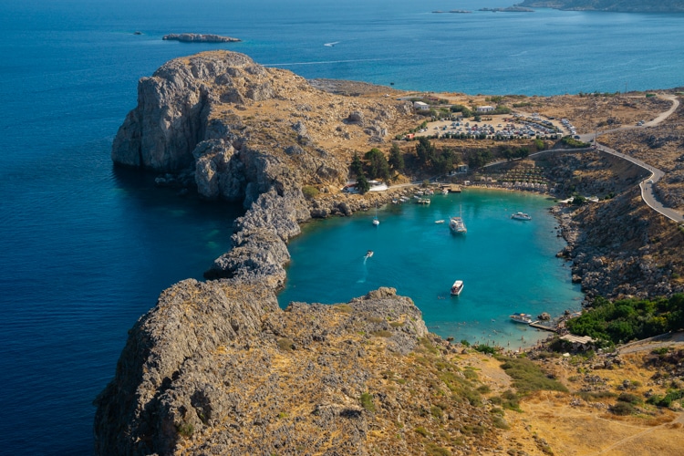Een luchtfoto van de schilderachtige baai bij de Akropolis van Lindos, met helder turquoise water omringd door ruige rotsformaties en een smal strand. Enkele boten dobberen in de kalme zee, terwijl een kronkelende weg toegang biedt tot dit serene landschap.