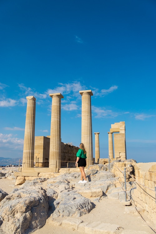 Een vrouw in een groene top en zwarte shorts staat op een rotsachtig pad bij de oude Dorische zuilen van de Akropolis van Lindos. De blauwe lucht en historische architectuur vormen een indrukwekkend contrast, terwijl de ruïnes de grandeur van het verleden benadrukken.
