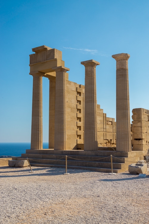 Een close-up van de goed bewaarde Dorische tempelzuilen op de Akropolis van Lindos, met een helderblauwe lucht en de Egeïsche Zee op de achtergrond. De symmetrische architectuur en historische details benadrukken de klassieke schoonheid van de Griekse oudheid.