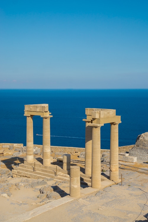 Een uitzicht op de overblijfselen van een oude tempel op de Akropolis van Lindos, met twee gerestaureerde Dorische zuilen en een helderblauwe zee op de achtergrond. Het minimalistische ontwerp van de tempel benadrukt de harmonie tussen architectuur en natuur.