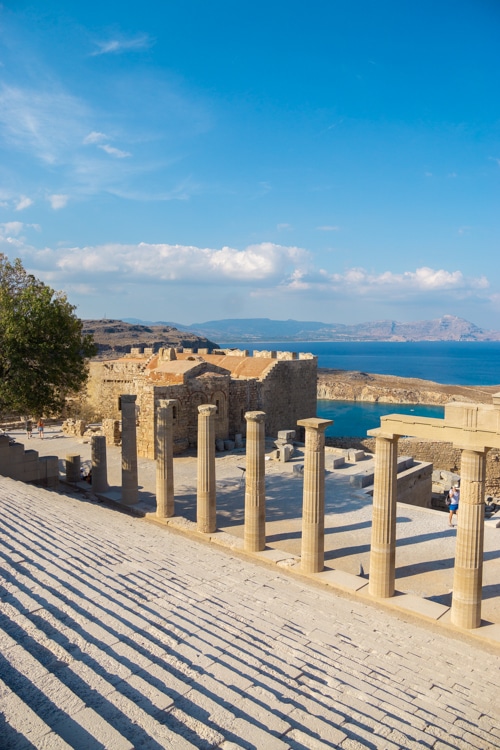 Een indrukwekkend uitzicht op de Akropolis van Lindos, met een rij Dorische zuilen en een trap die naar de oude ruïnes leidt. De zee en omliggende heuvels creëren een schilderachtige achtergrond, terwijl het zonlicht de schaduwen van de zuilen benadrukt.
