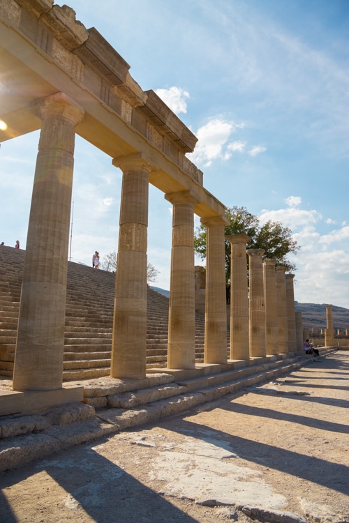 Een close-up van de Dorische zuilengalerij op de Akropolis van Lindos, met zonnestralen die door de openingen vallen. De perfect uitgelijnde zuilen en het spel van licht en schaduw benadrukken de grootsheid van de oude Griekse architectuur.