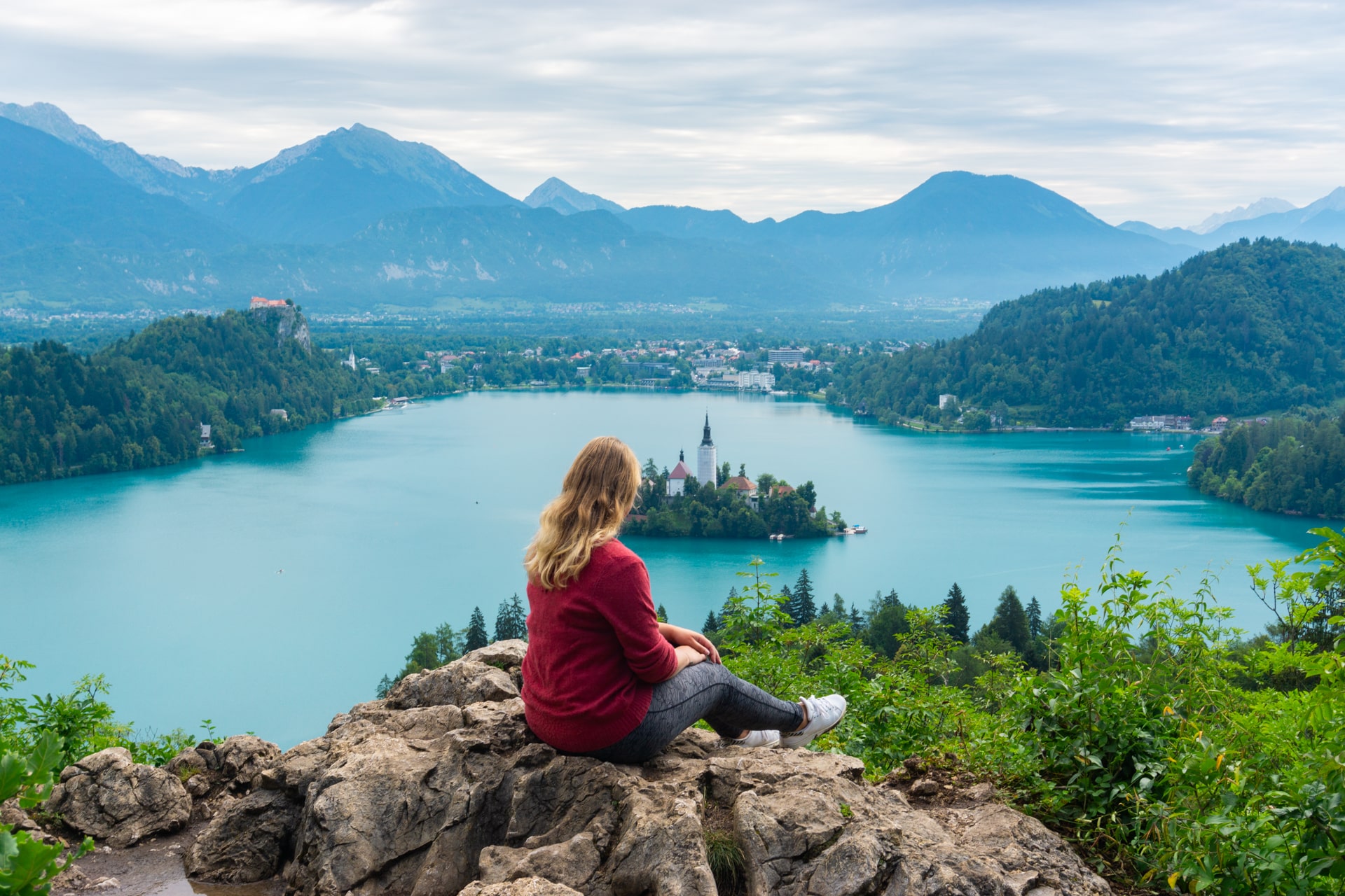 Een vrouw geniet in haar eentje van het uitzicht vanaf een rotsachtige uitkijkpunt over het turquoise meer van Bled in Slovenië, met het beroemde eiland Bled en de kerk centraal in beeld. Omringd door groene heuvels en bergen onder een bewolkte hemel, is dit een perfecte bestemming voor soloreizen en serene natuurbeleving.